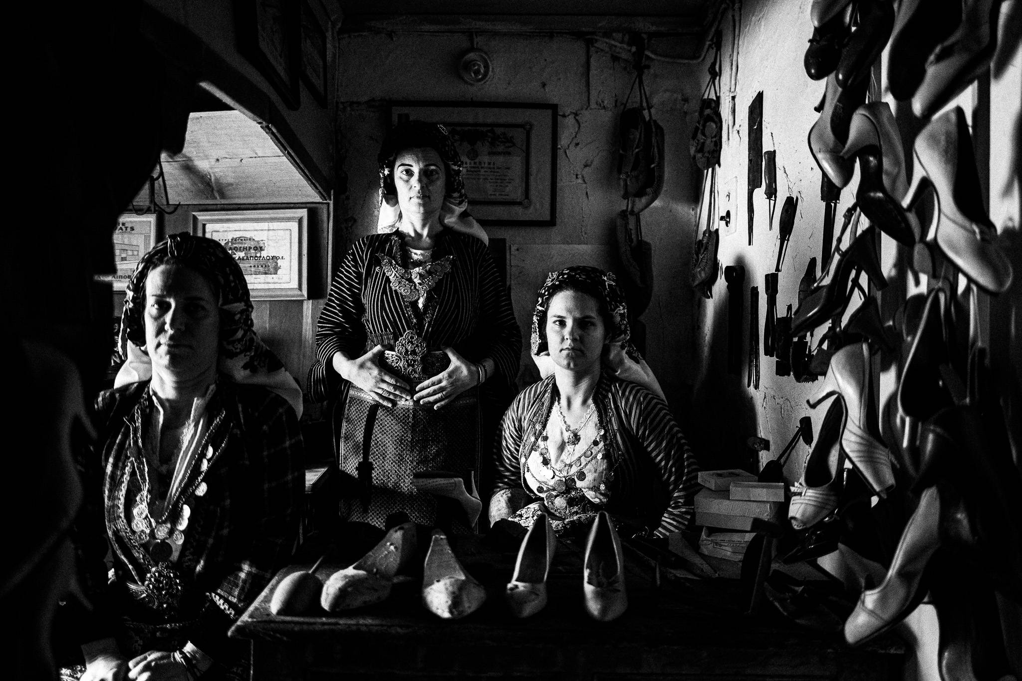 Black and white photo of three women in traditional plaid costumes in an old cobbler’s workshop. Vintage shoe forms, tools, and a Singer sewing machine surround them, illuminated by window light, capturing a timeless craft environment.