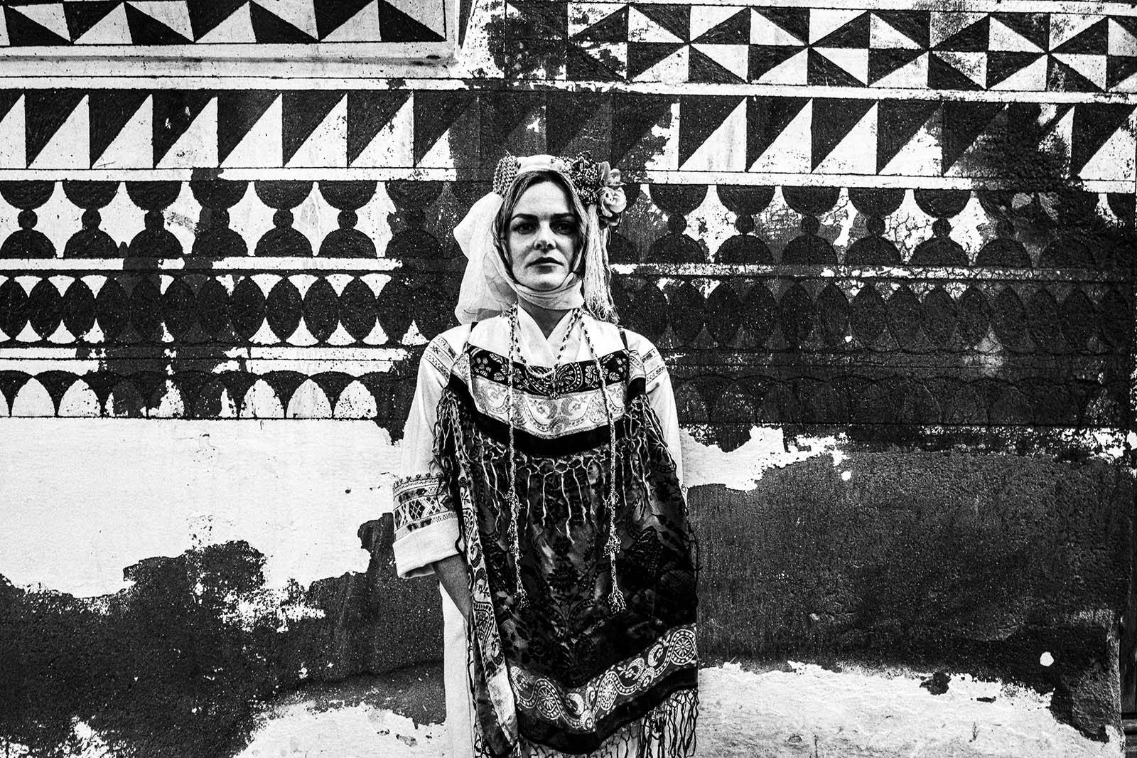 Black-and-white portrait of a woman wearing traditional costume in front of the geometric wall of Pyrgi, Chios.