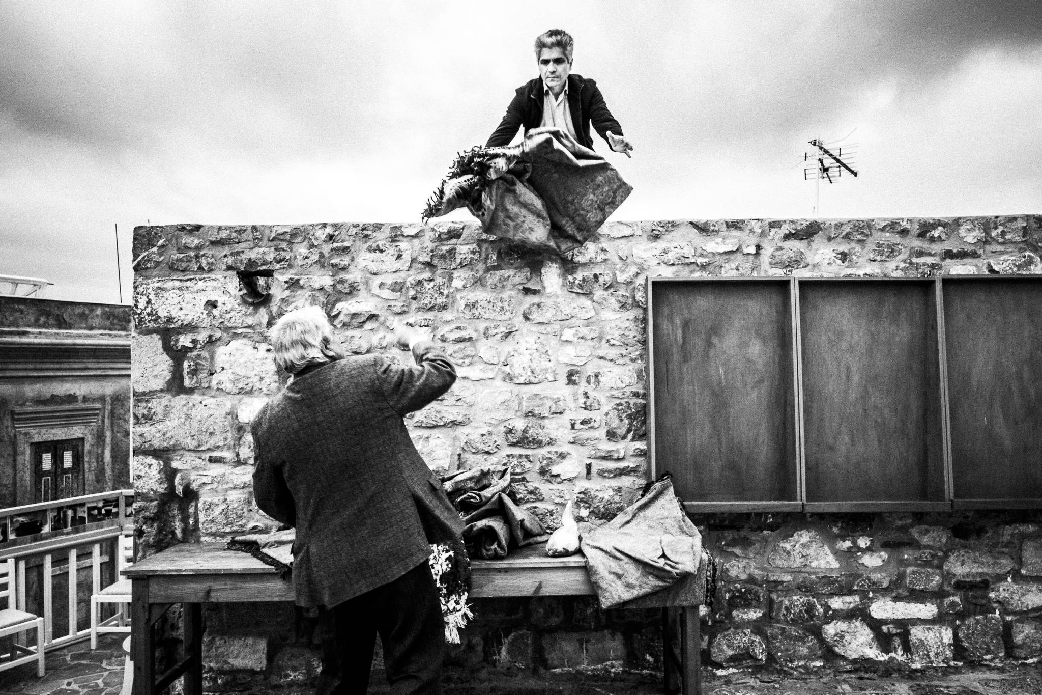 A man trowing a tapestry to another, standing on a roof. Olympos, Karpathos, Greece. 