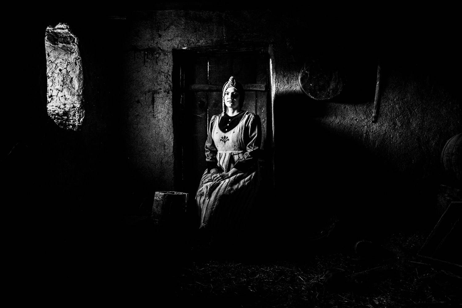 Sitting portrait of a woman in an old warehouse, wearing the local traditional dress. Lefki, Kastoria, Macedonia, Greece. Black and white photograph by George Tatakis