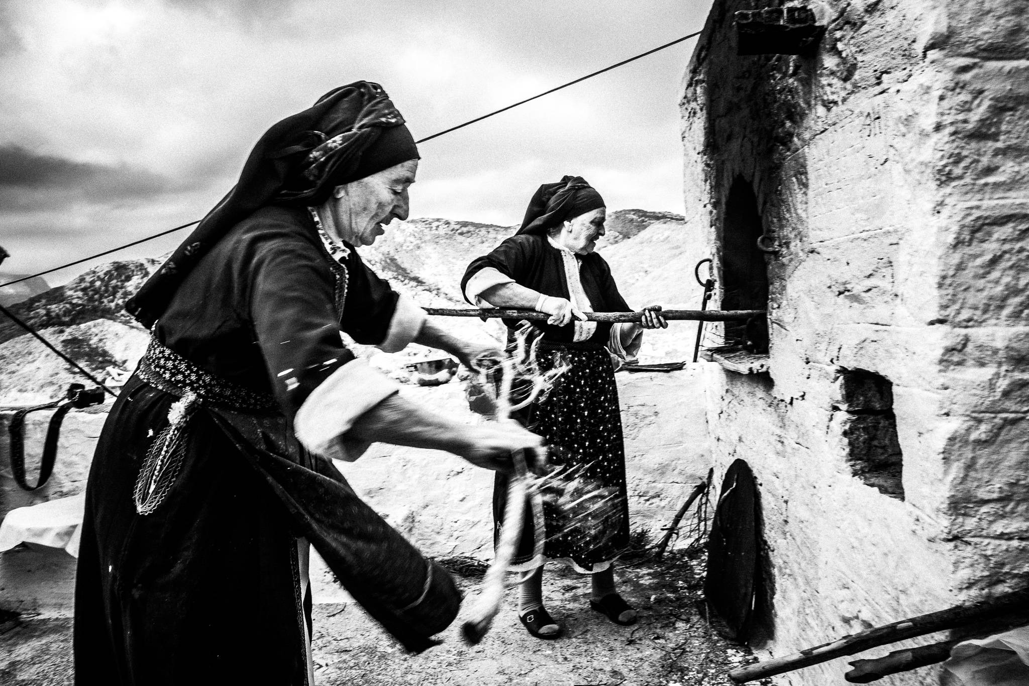 Two women in the traditional local costumes, lighting up the wood-burning oven. Olympos, Karpathos, Greece
