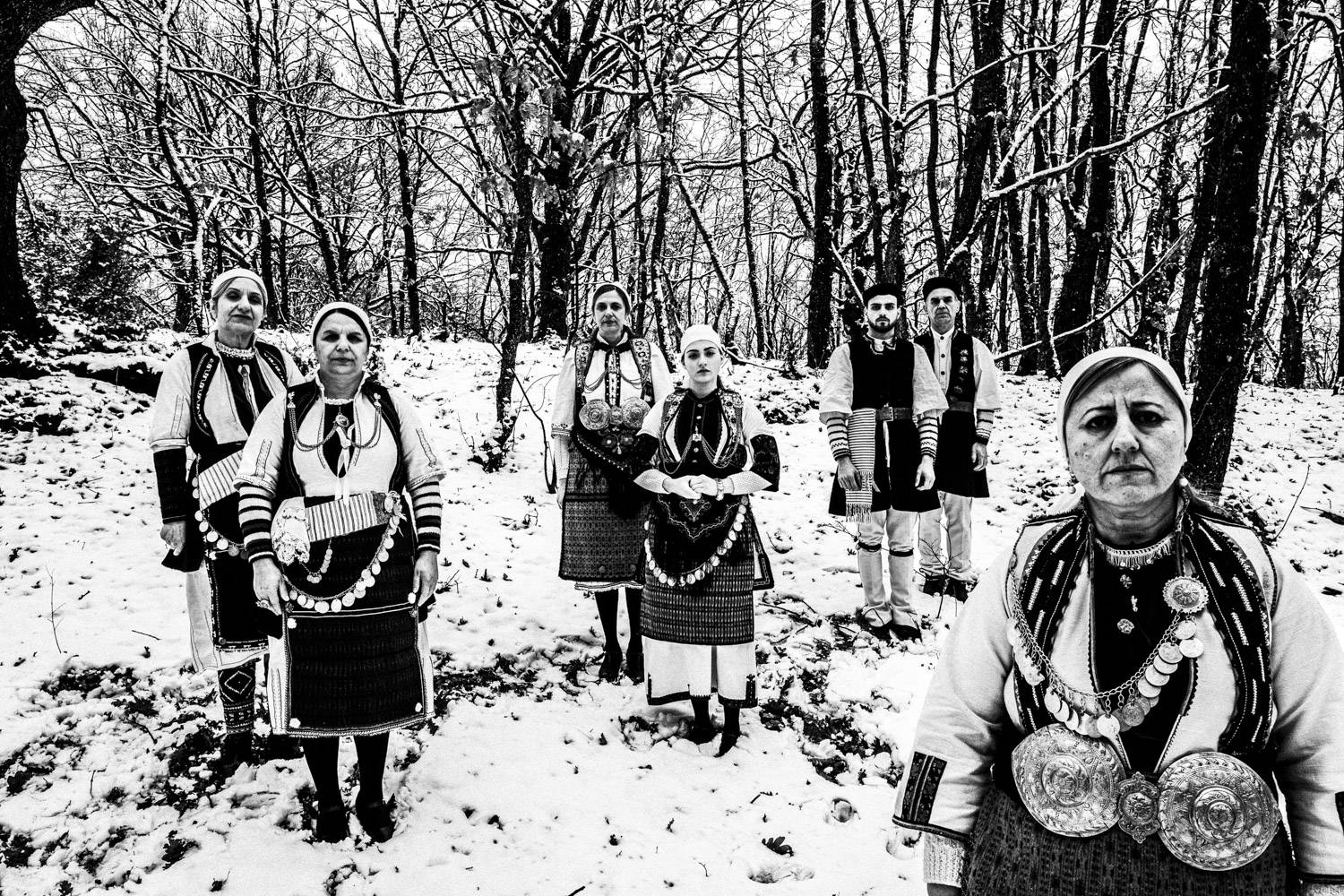 Seven persons posing at a forest covered in snow, wearing the local traditional costumes. Kladorachi, Florina, Macedonia, Greece. Black and white photograph by George Tatakis