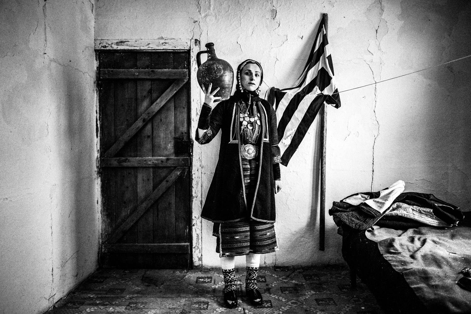 A woman hoding an urn next to the Greek flag, in Volax, Macedonia, Greece. Black and white photograph by George Tatakis