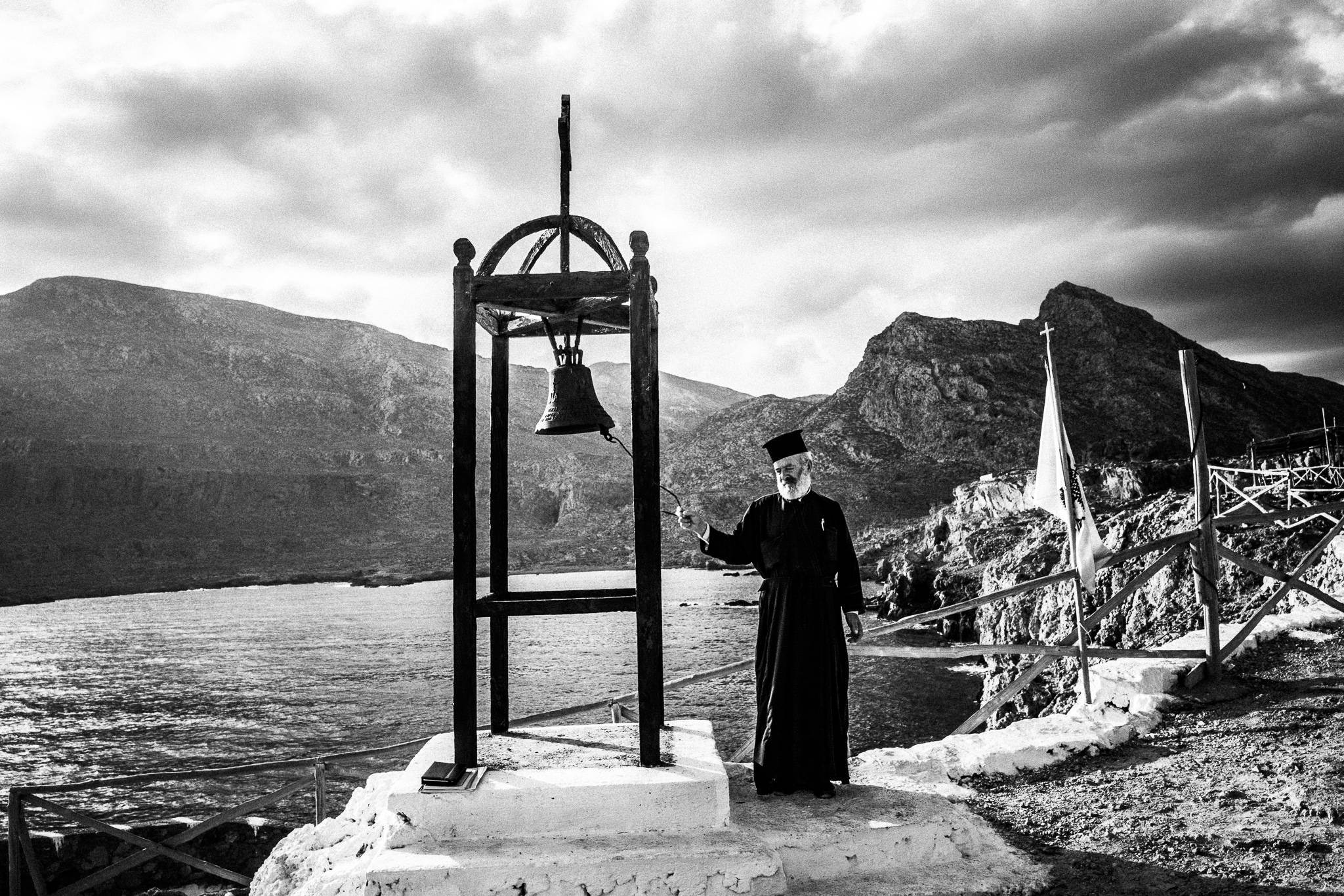 Striking the bell to call the faithful to the morning liturgy at the site of the ancient city of Brykous, Olympos, Karpathos, Greece. Black and white photography by George Tatakis