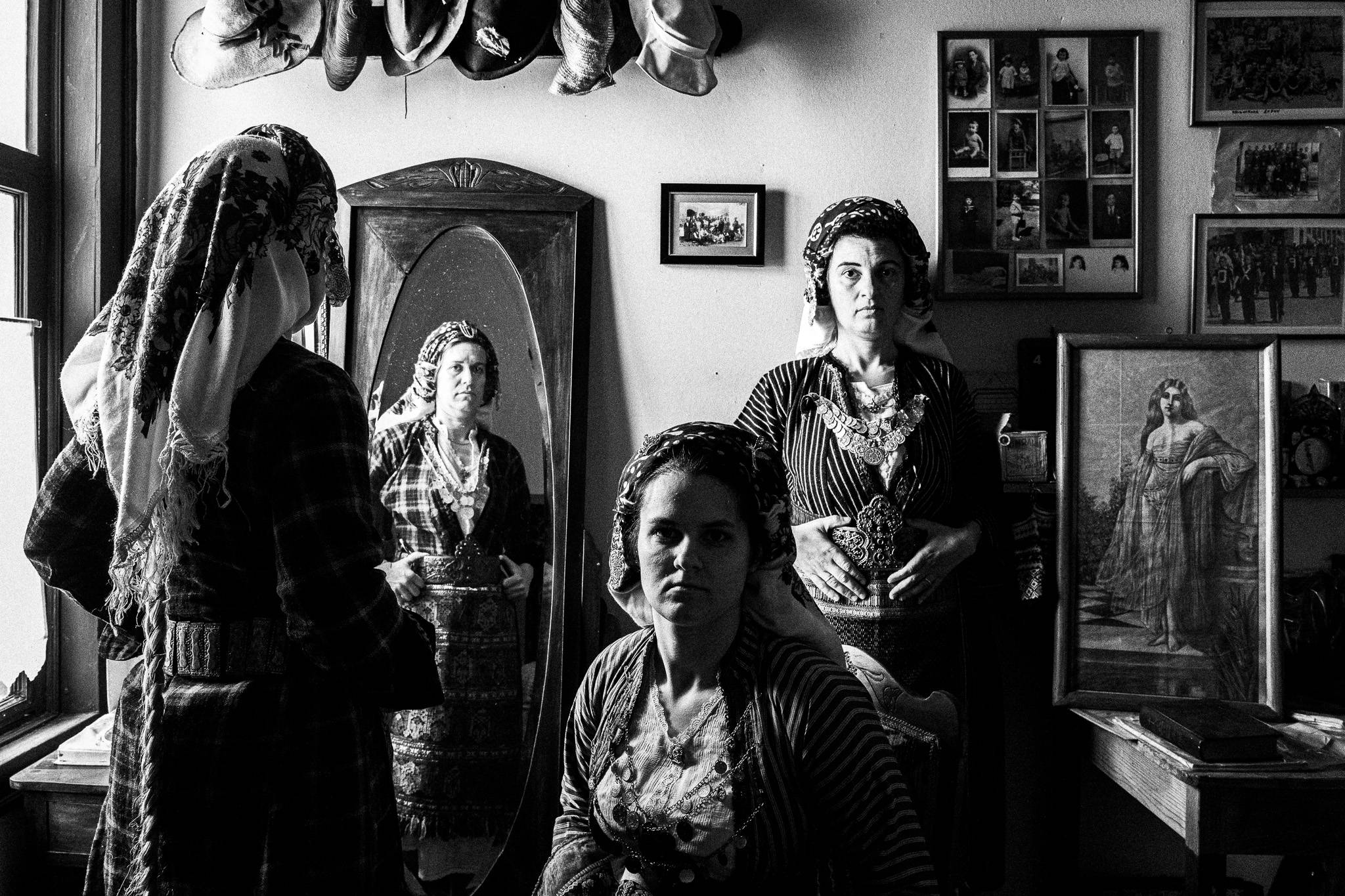 Black and white photo of three Soufli women in traditional dress inside a historic room. One woman’s reflection appears in a tall mirror as another stands beside it; old photographs and artwork decorate the walls, highlighting the reflection of past and present.