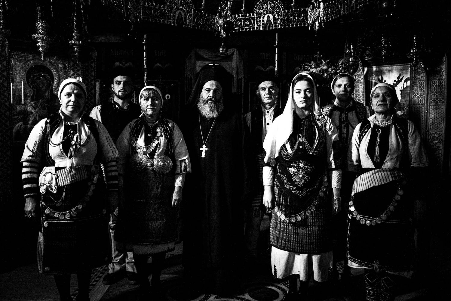A priest with a group of people in their traditional clothes, inside the church of the local Monastery. Kladorachi, Florina, Macedonia, Greece. Black and white photograph by George Tatakis