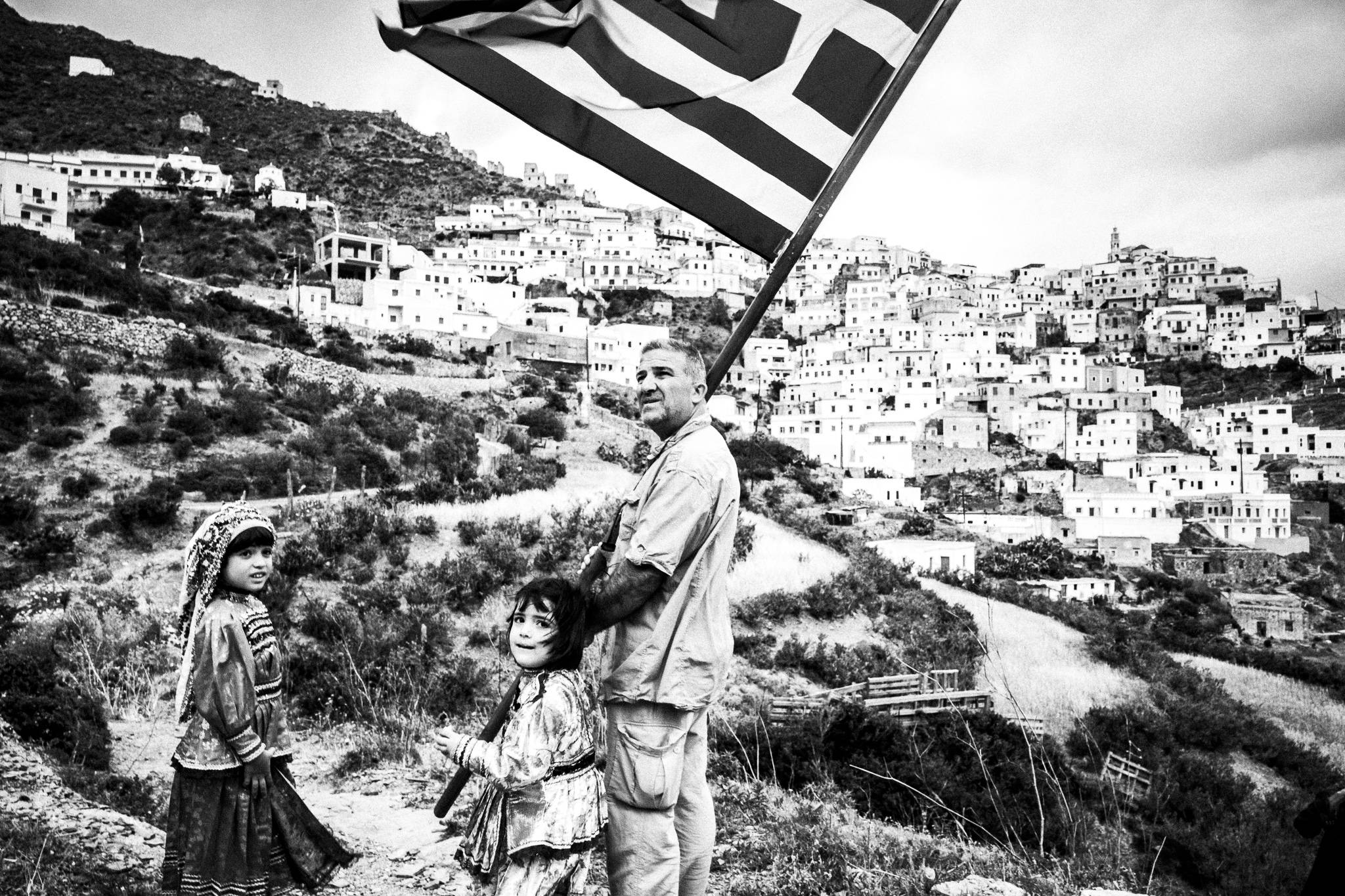 A man with his two young daughters, holding the Greek flag, overlooking the village. Olympos, Karpathos, Greece. 