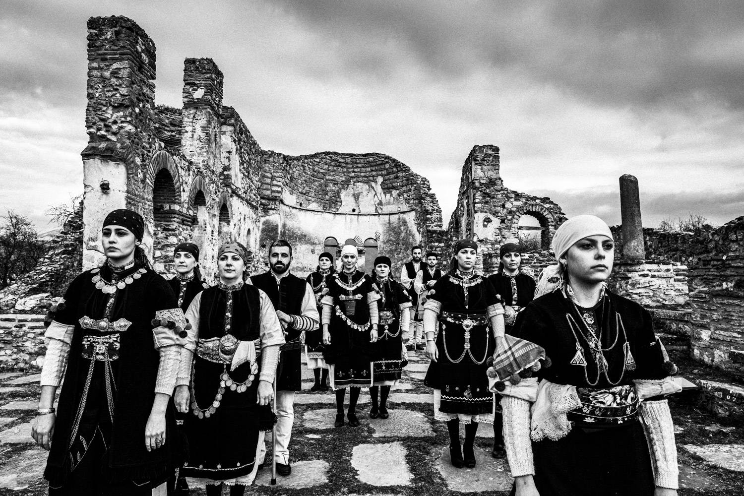 A group of 13 persons posing at the Cathedral of Saint Achilles, wearing the traditional local attire. Prespes Lakes, Macedonia, Greece. Black and white photograph by George Tatakis