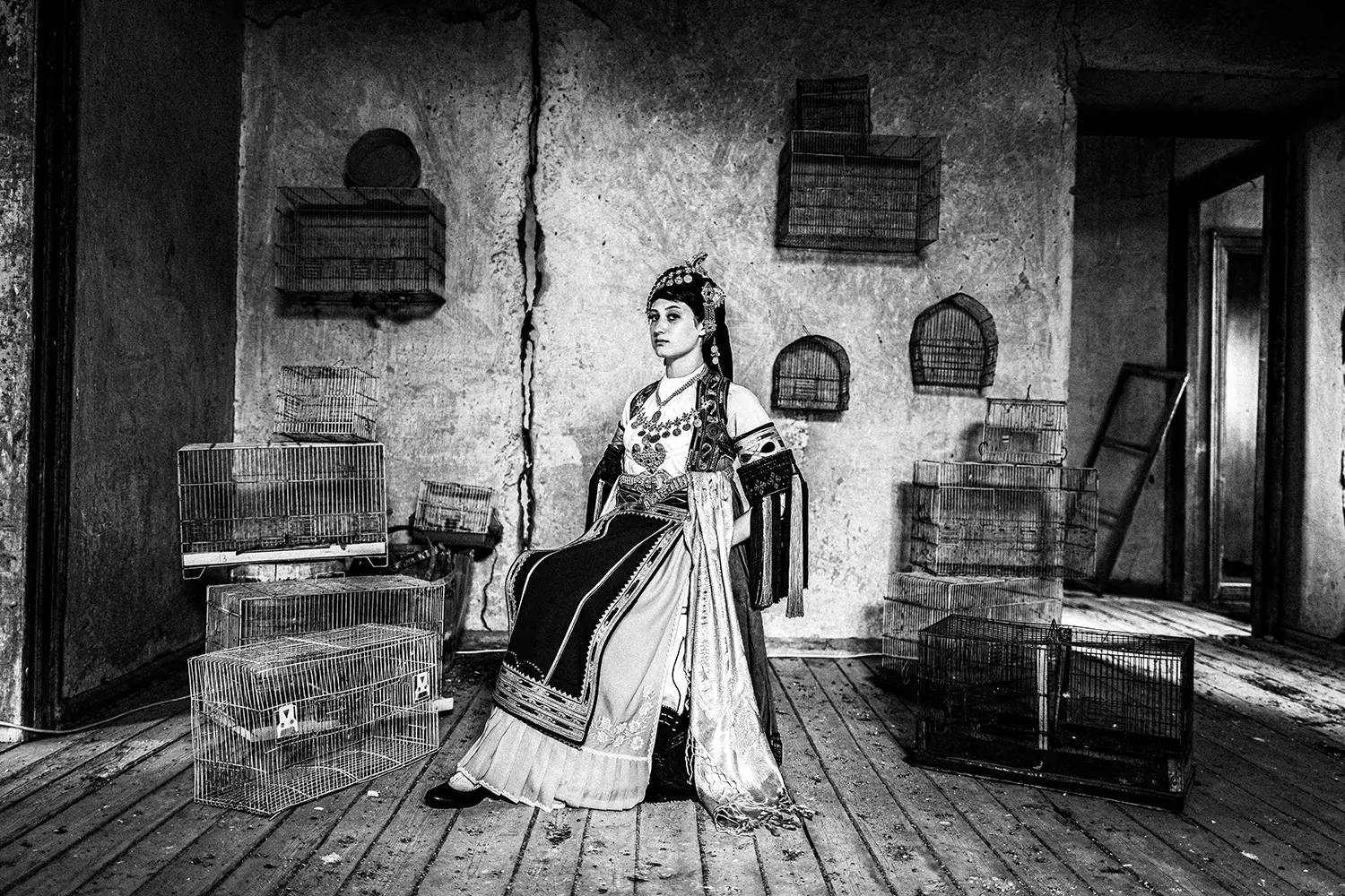 A woman inside a room full of bird cages, at a mansion in Glinos, wearing the traditional Karagouna dress. Trikala, Thessaly, Greece. Black and white photography by George Tatakis