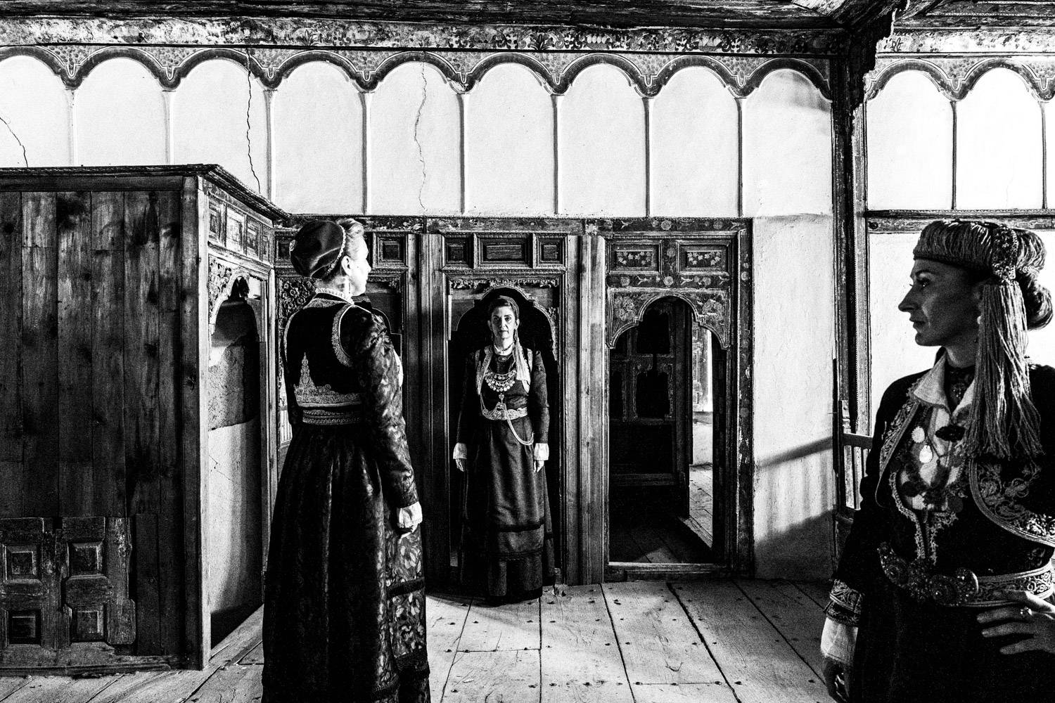 Three women standing inside a Mansion in Siatista, wearing the urban dresses of Kozani, Macedonia, Greece. Black and white photograph by George Tatakis