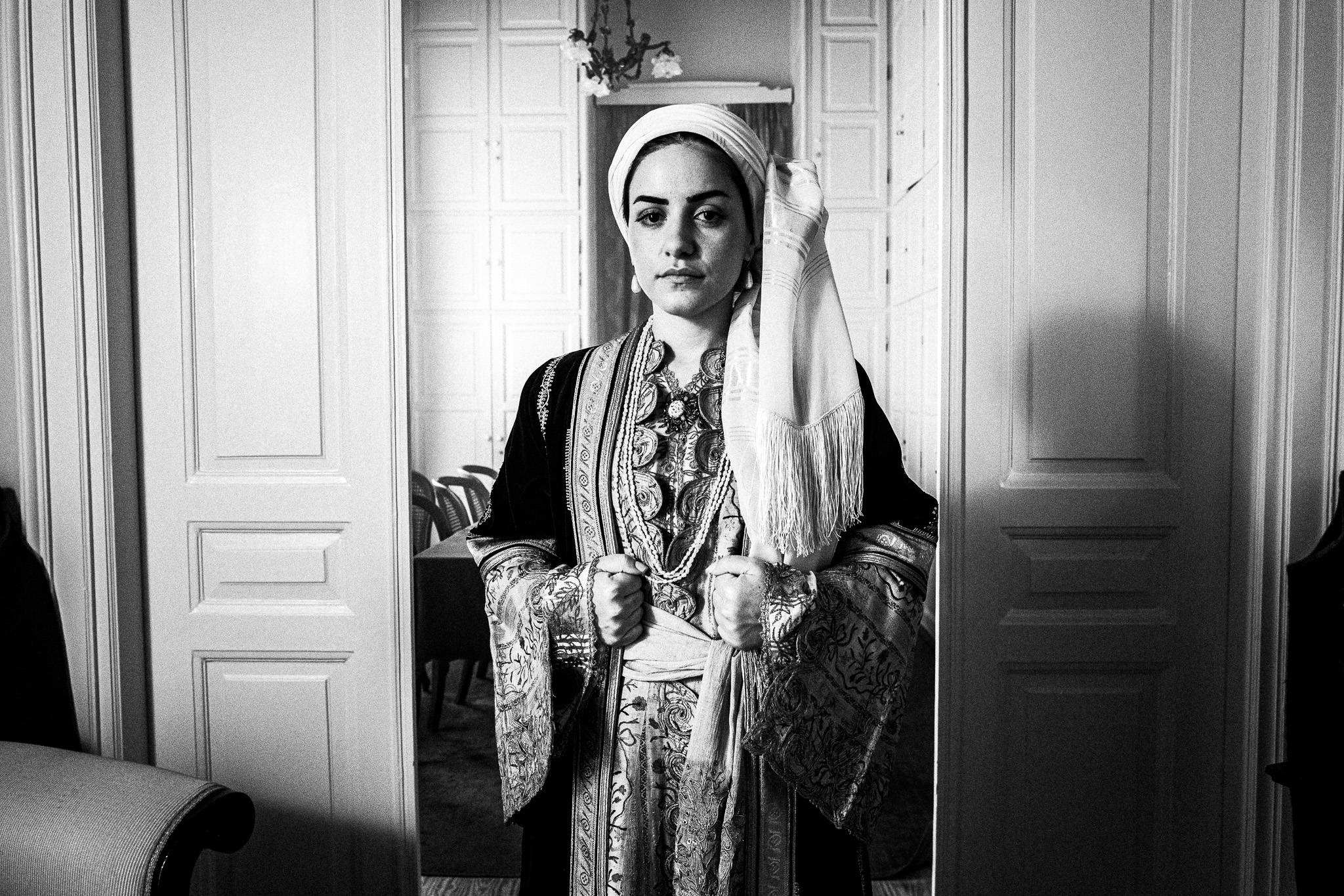 Woman in traditional urban attire from Kalamata stands in a doorway, holding fabric. Ornate patterns on clothing; backdrop shows a room with chairs. Black and white photo.
