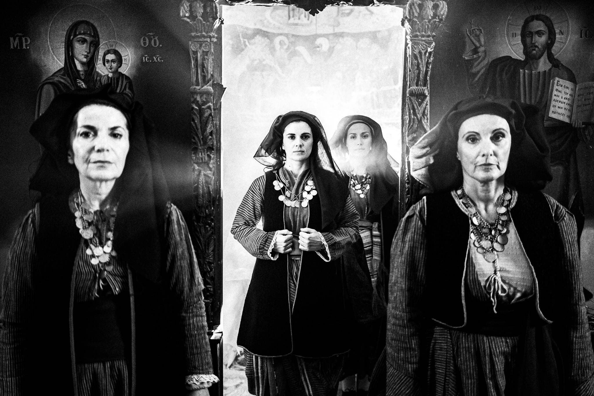 Three women in traditional attire of Alagonia at the monastery of Mandraki, framed by religious icons. The setting is dimly lit, creating a somber mood.