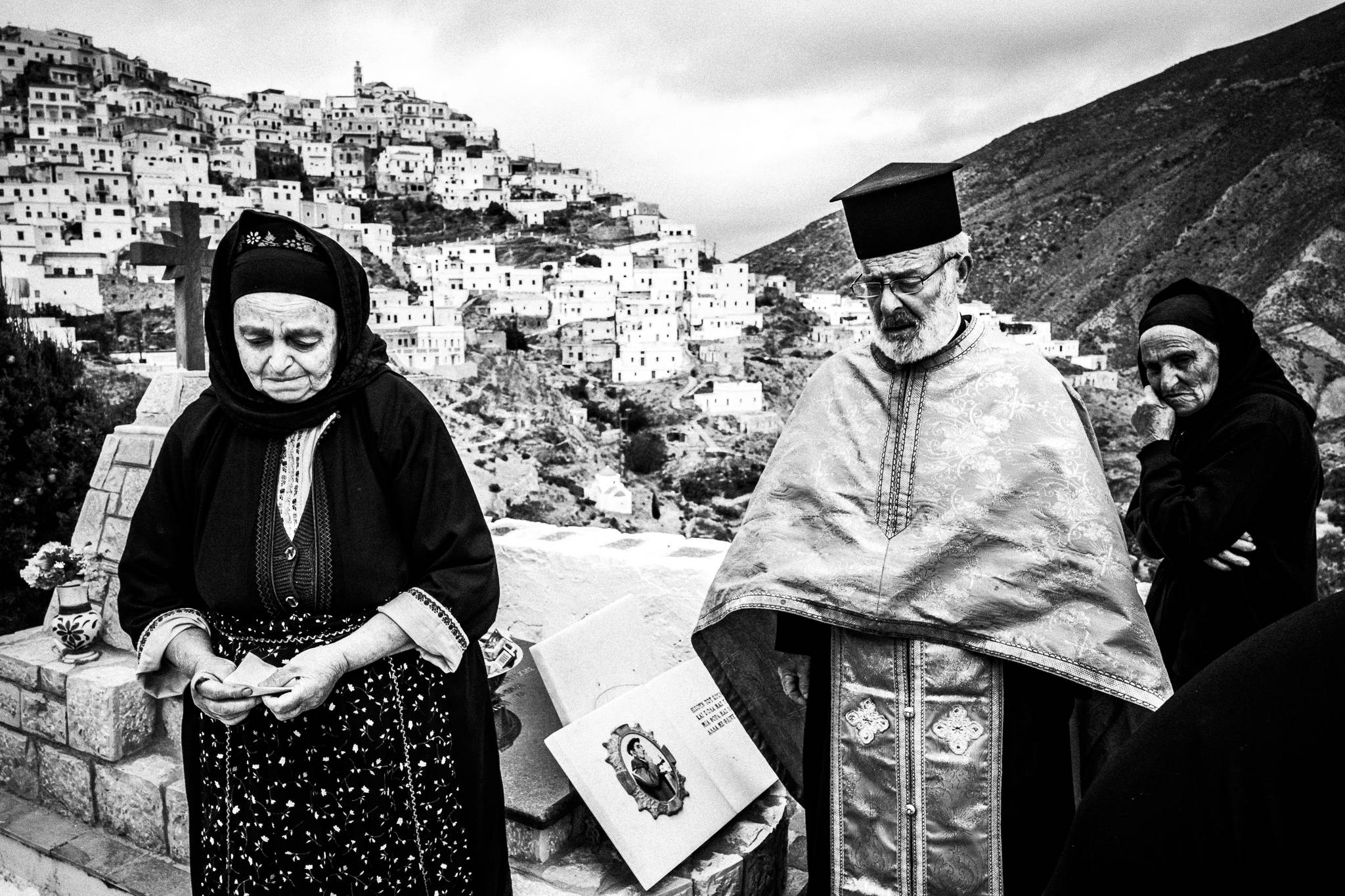 The priest with two local women in their traditional attire, performing a memorial service at the local cemetery, overlooking the village. Olympos, Karpathos, Greece. 