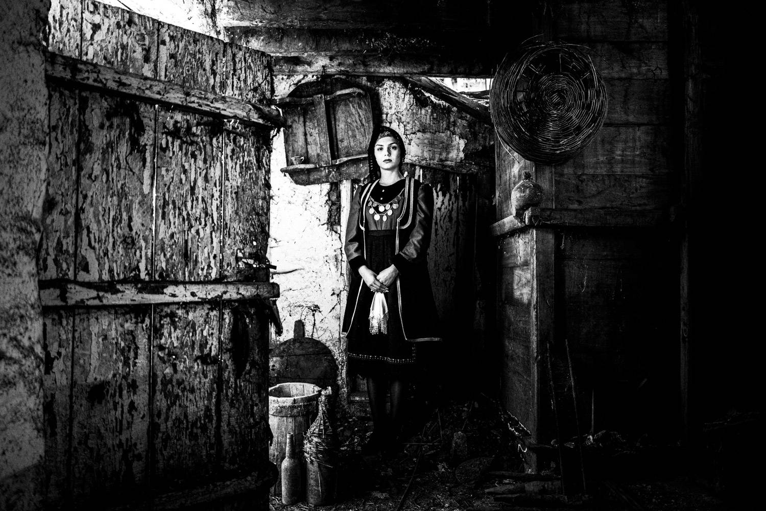 A woman posing inside an old house, wearing the local folklore dress. Lefki, Kastoria, Macedonia, Greece. Black and white photograph by George Tatakis