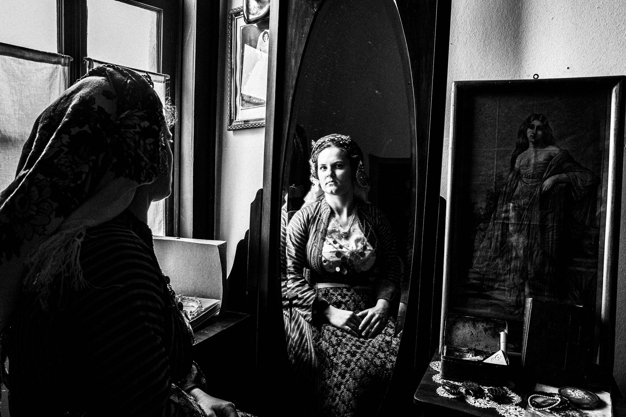 Black and white close-up portrait of a young woman in Soufli traditional attire gazing into an antique mirror. Her face is illuminated as she meets the camera, with her embroidered headscarf and coin necklace visible, conveying a reflection on cultural identity.