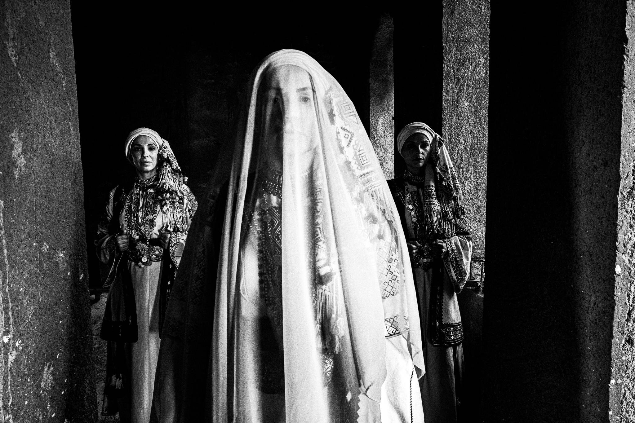 Three women in traditional attire of Kyparissia stand in dim light. The central woman is veiled. The setting is inside a local house. Peloponnese, Greece