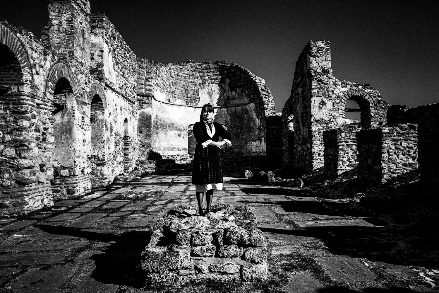 A woman posing at the center of the ruined Cathedral of Saint Achilles, wearing the local attire. Prespes Lakes, Macedonia, Greece. Black and white photograph by George Tatakis