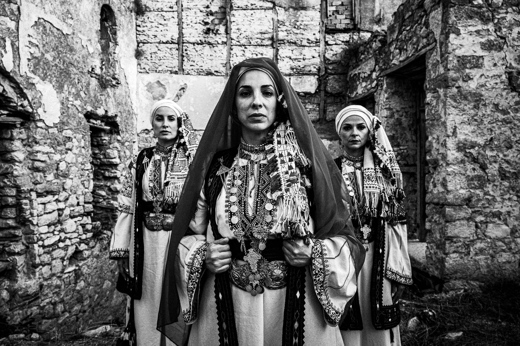 Three women in traditional attire of Kyparissia, stand solemnly in a weathered stone ruin. Ornate patterns and jewelry adorn their outfits.