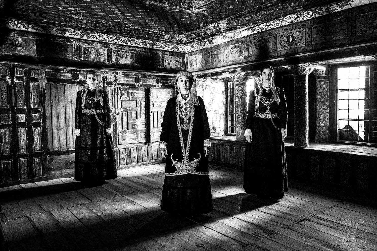 Three women standing inside a Mansion in Siatista, wearing the local costumes of Kozani, Macedonia, Greece. Black and white photograph by George Tatakis