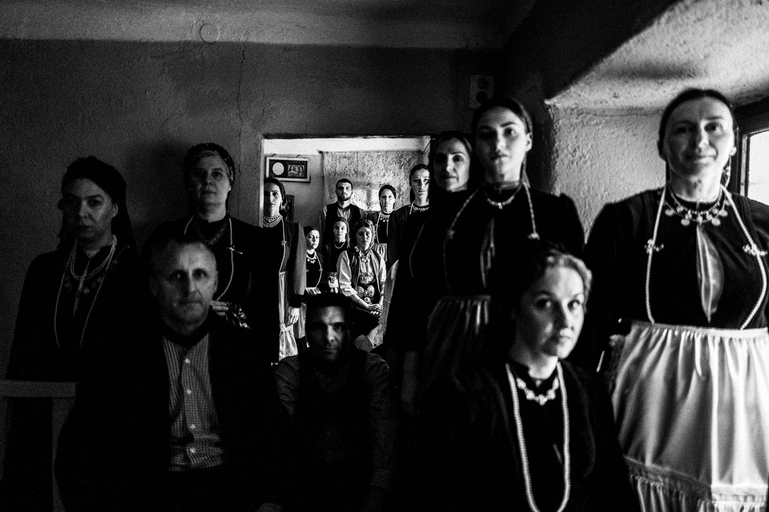 A group of 15 persons inside a small local traditional home, wearing the local folklore attire. Polypotamos, Florina, Macedonia, Greece. Black and white photograph by George Tatakis