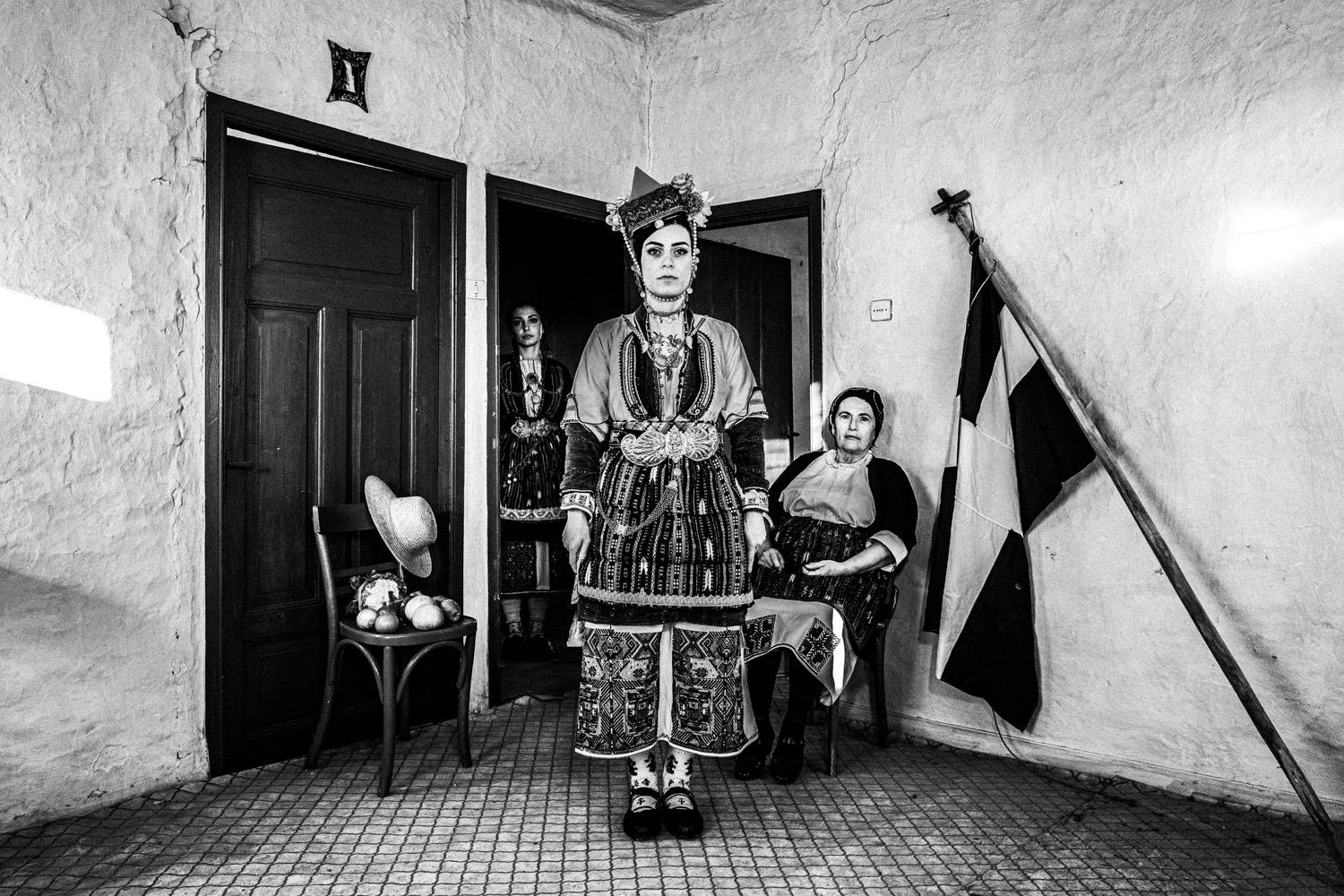 Three women at a local traditional home, in their traditional clothes, next to the Greek flag. Aggelochori, Naoussa, Macedonia, Greece. Black and white photograph by George Tatakis