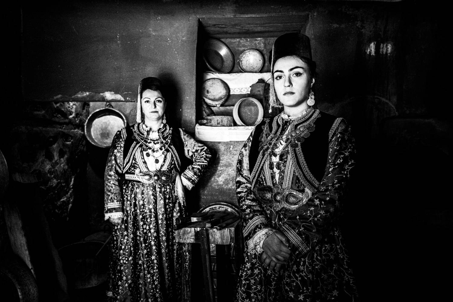 Mother and daughter in the local traditional attire, posing inside the kitchenette of a traditional Mansion. Kastoria, Macedonia, Greece. Black and white photograph by George Tatakis