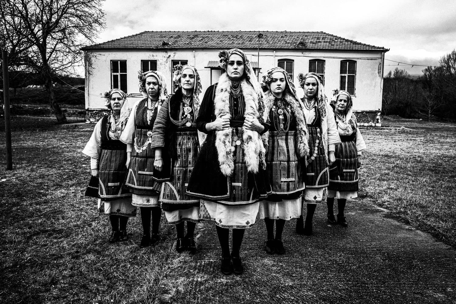 A group of seven women outside the local school, posing in an arrow formation in their local folklore dresses. Skopos, Florina, Macedonia, Greece. Black and white photograph by George Tatakis