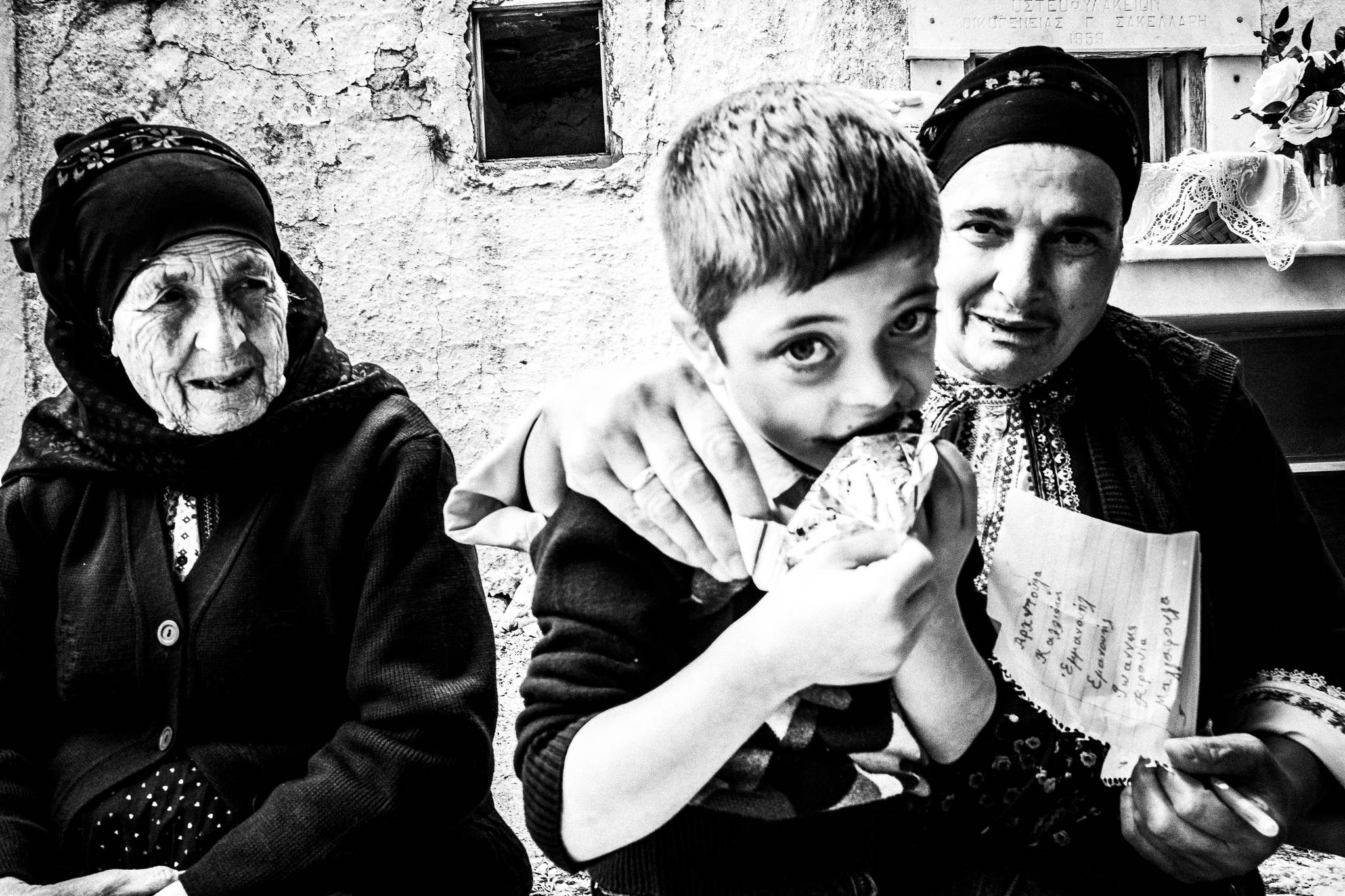 A kid eating candy with two women in traditional clothes, at the local cemetery. Olympos, Karpathos, Greece. 