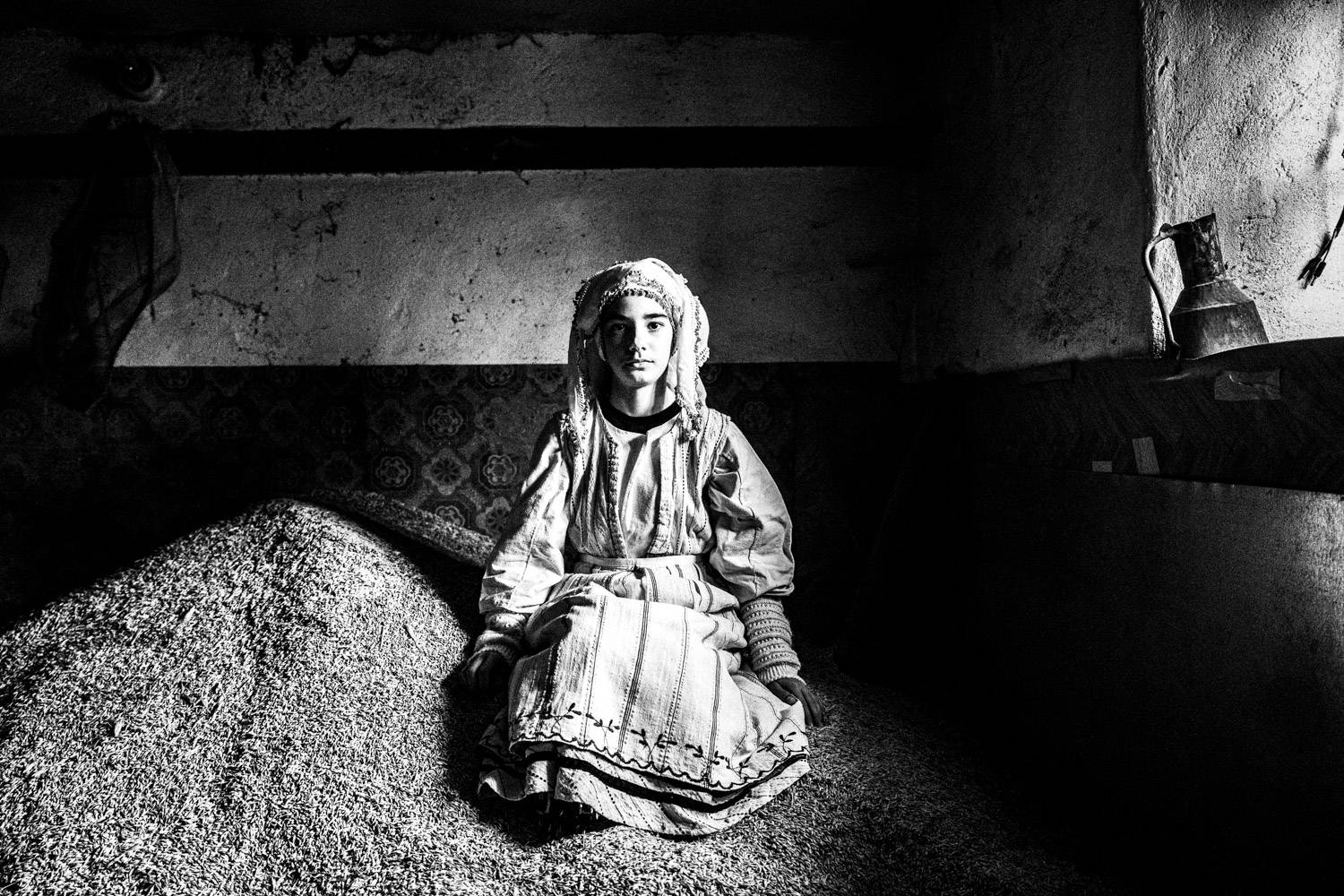 Portrait of a girl, wearing the local folklore dress, sitting on a pile of grain at an abandoned house. Ermakia, Ptolemaida, Macedonia, Greece. Black and white photograph by George Tatakis