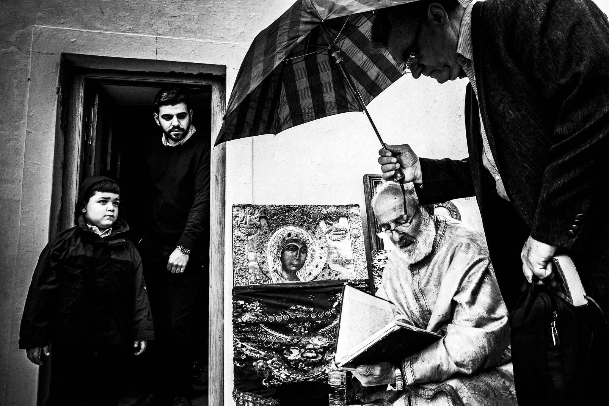 A man holding an umbrella for the local priest, outside a church. Olympos, Karpathos, Greece. 