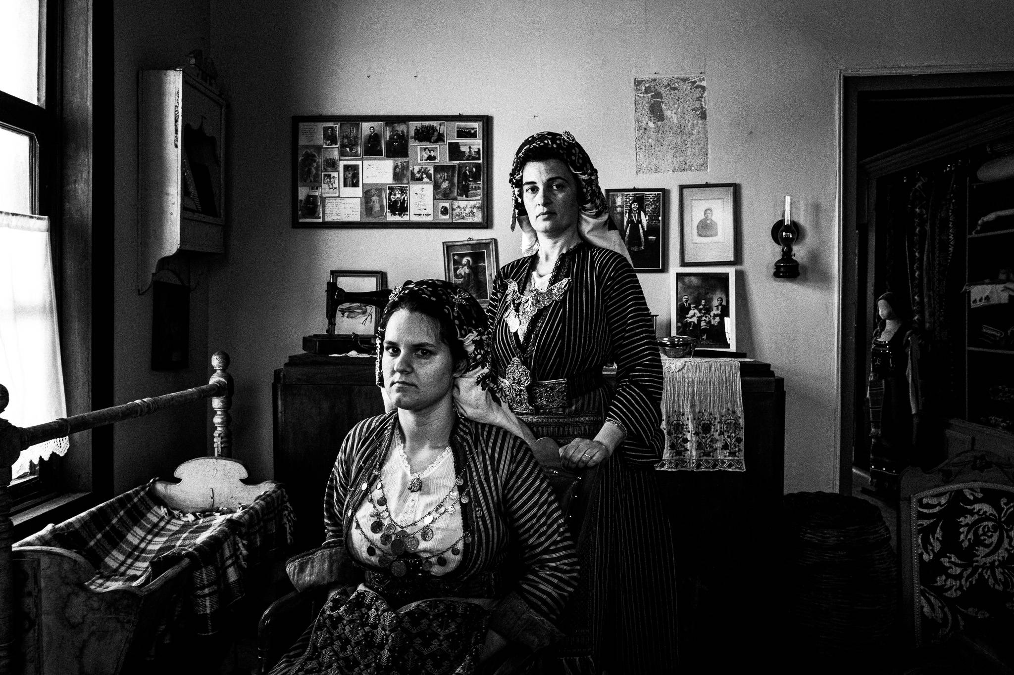 Black and white photo of two women in traditional Soufli costumes sitting and standing in an old-fashioned room with family photos on the wall and a wooden cradle, symbolizing heritage preserved in a home setting.