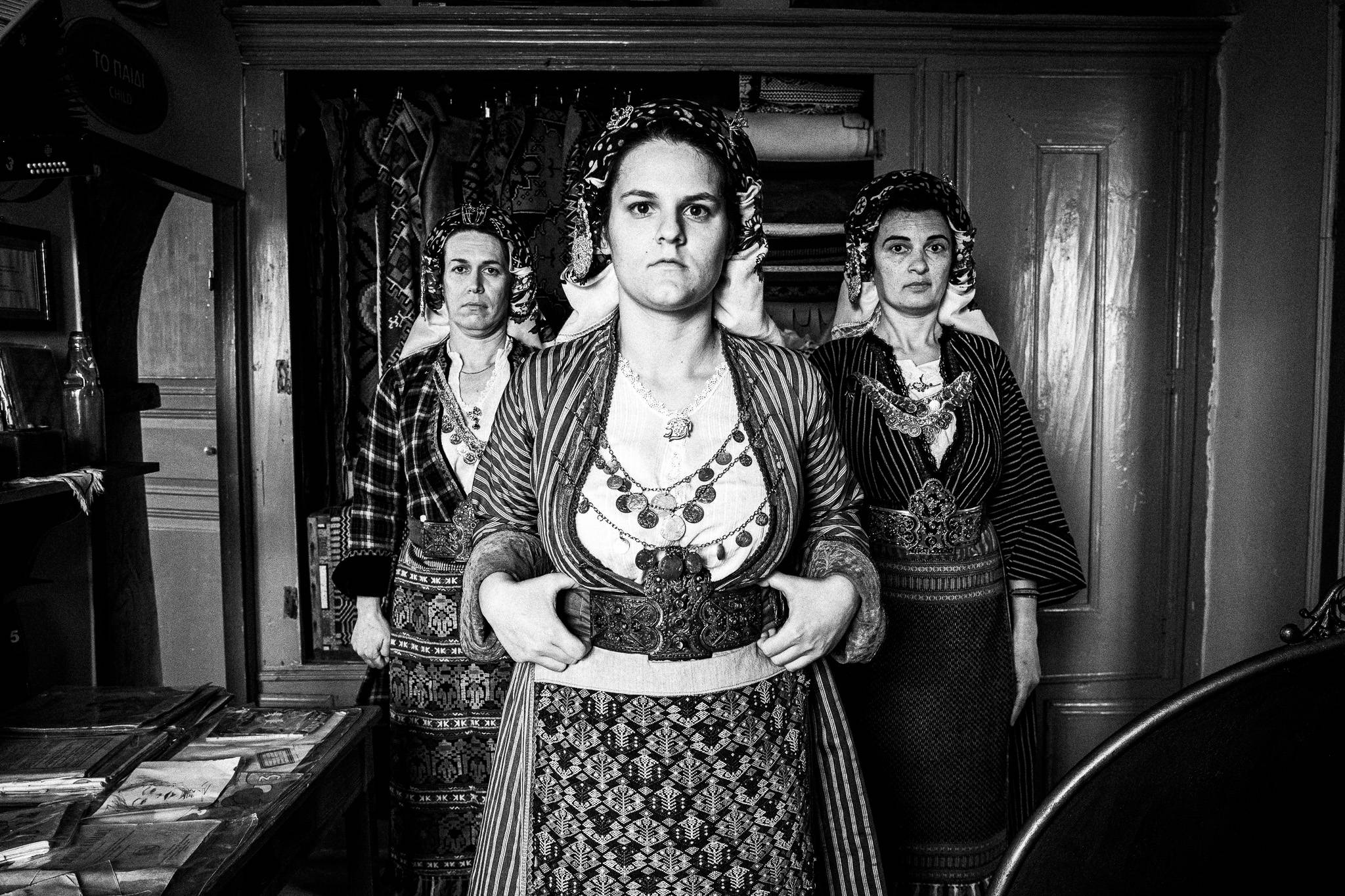 Black and white photograph of three Soufli women standing shoulder to shoulder in full traditional costume against a doorway backdrop. They face the camera with proud expressions, showcasing detailed textiles, belts, and jewelry of Thracian folk dress, representing the guardians of local tradition.