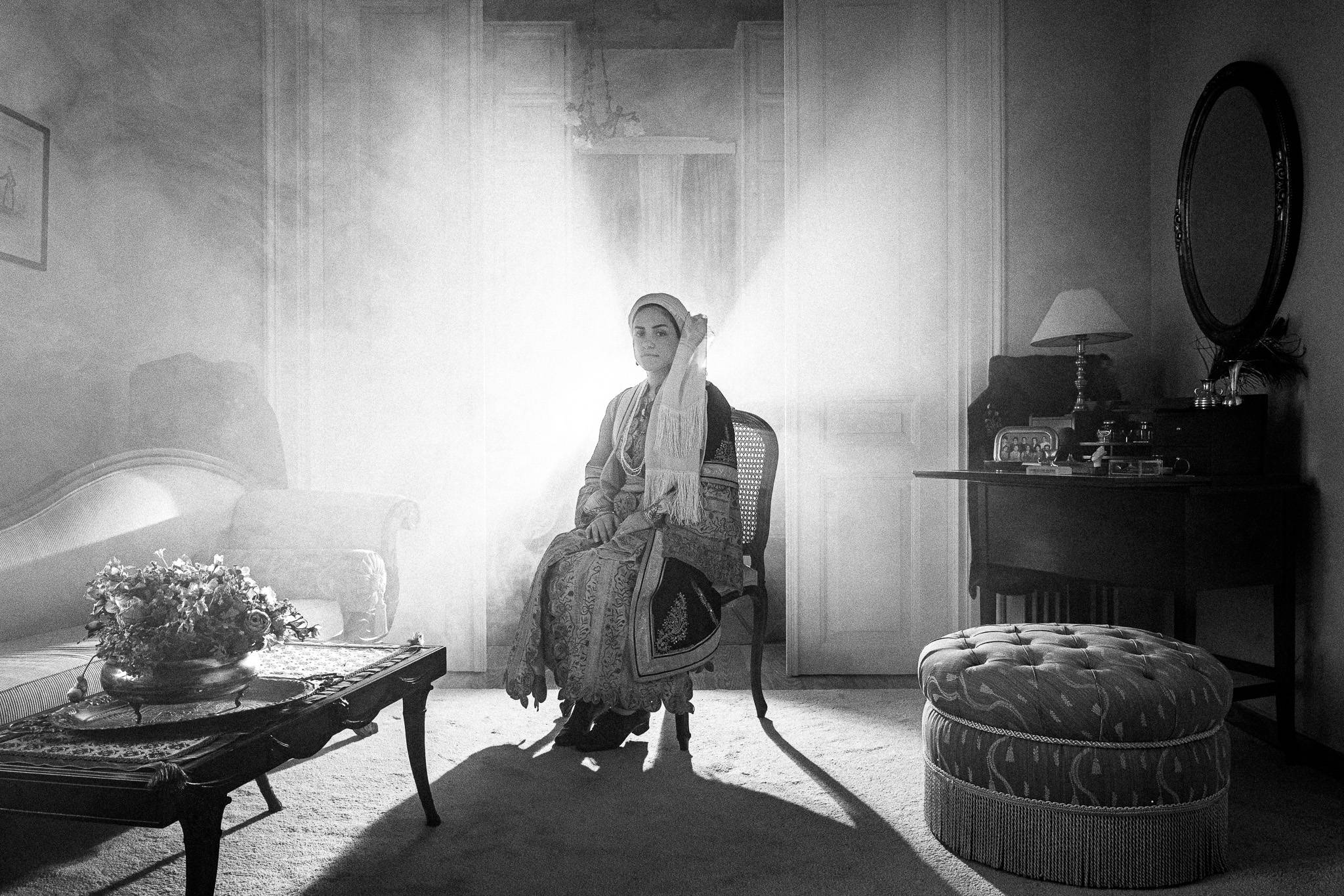 A woman in traditional urban attire of Kalamata sits on a chair in a dim, vintage room with elegant furniture and floral decor; light streams dramatically behind.