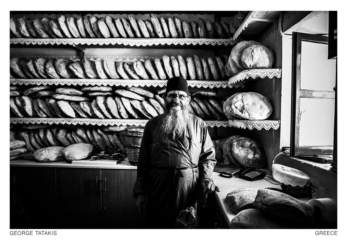 The priest is standing in front of prepared bread, for the celebration of St Paraskevi on the island of Amorgos, Cyclades, Greece. Black and white photography postcard by George Tatakis, project Ethos.