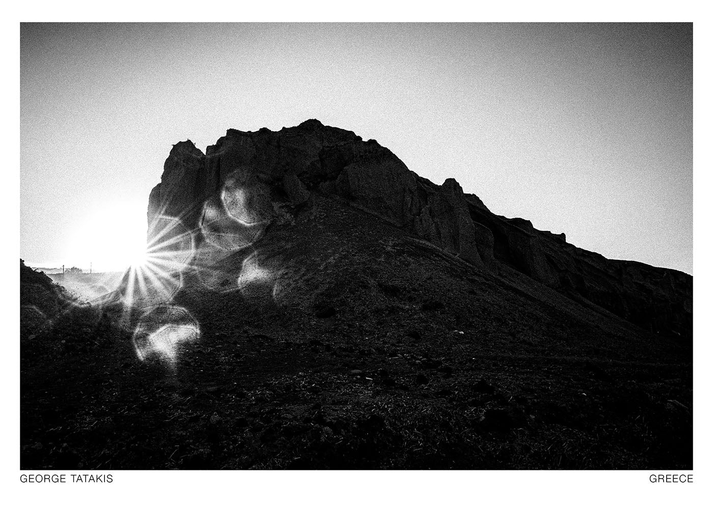 A volcanic boulder against the sun at a cycladic beach. Black and white photography postcard by George Tatakis, project Chorōs.